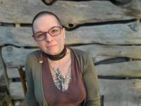 A non-binary femme person with red glasses, a tattoo of a pine tree, beaded earrings and a leather collar smiles warmly against a background of a log wall.