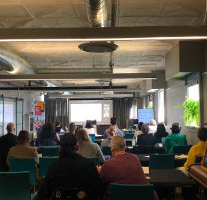 A photo from the back of the room displays the Reimagining Livelihoods Forum audience, their bodies turned away from the camera. The screen at the front of the room is hard to see, but displays the online audience and presenters.