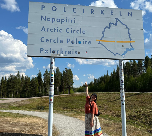 A photo of Maegan Ellis standing under and pointing at a sign for the arctic circle. She is pictured with long brown hair under a ballcap, an orange skirt, and a traditional ribbon skirt.
