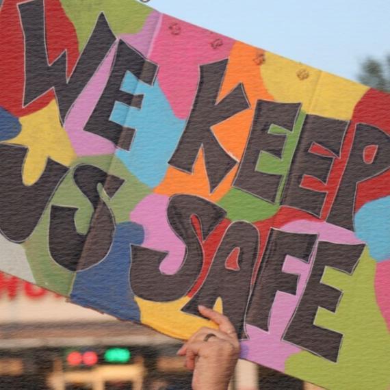 A hand holds a hand-painted cardboard sign that says “we keep us safe” in black block letters against a multicoloured abstract painted background.