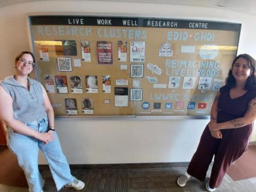 Composite Stories authors, Jordyne Craig and Vanessa Bonham, standing for a photo in front of the Centre's announcement board. 