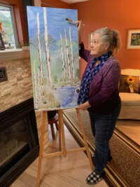 Image 2. An older white woman standing in a living room while painting at an easel. The painting is of a nature scene, with tall trees surrounded by water and the sky. There is a fireplace and a couch in the background. 