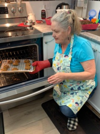 Image 4. An older white woman in a kitchen putting a tray of cookies in the oven. She is wearing a blue shirt and has an apron and oven mitt on.  