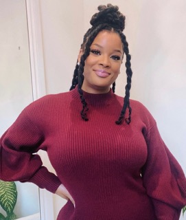 Tamara is pictured smiling with her left hand on her hip in front of a clean beige background. She is wearing a maroon dress and has dark-toned skin. Her dark brown hair has been partially tied into a bun on top of her head, with two locks left out to frame her face.