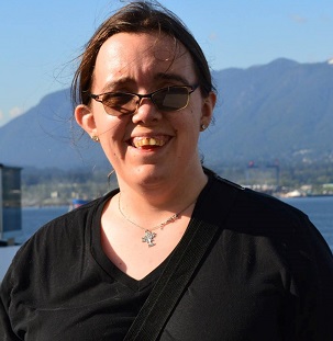 Maggie is pictured smiling in front of a scenic view. Their brown hair is tied back, and the sun is shining down on their light-toned skin. They are wearing sunglasses and a black T-shirt, with a silver necklace around their neck.